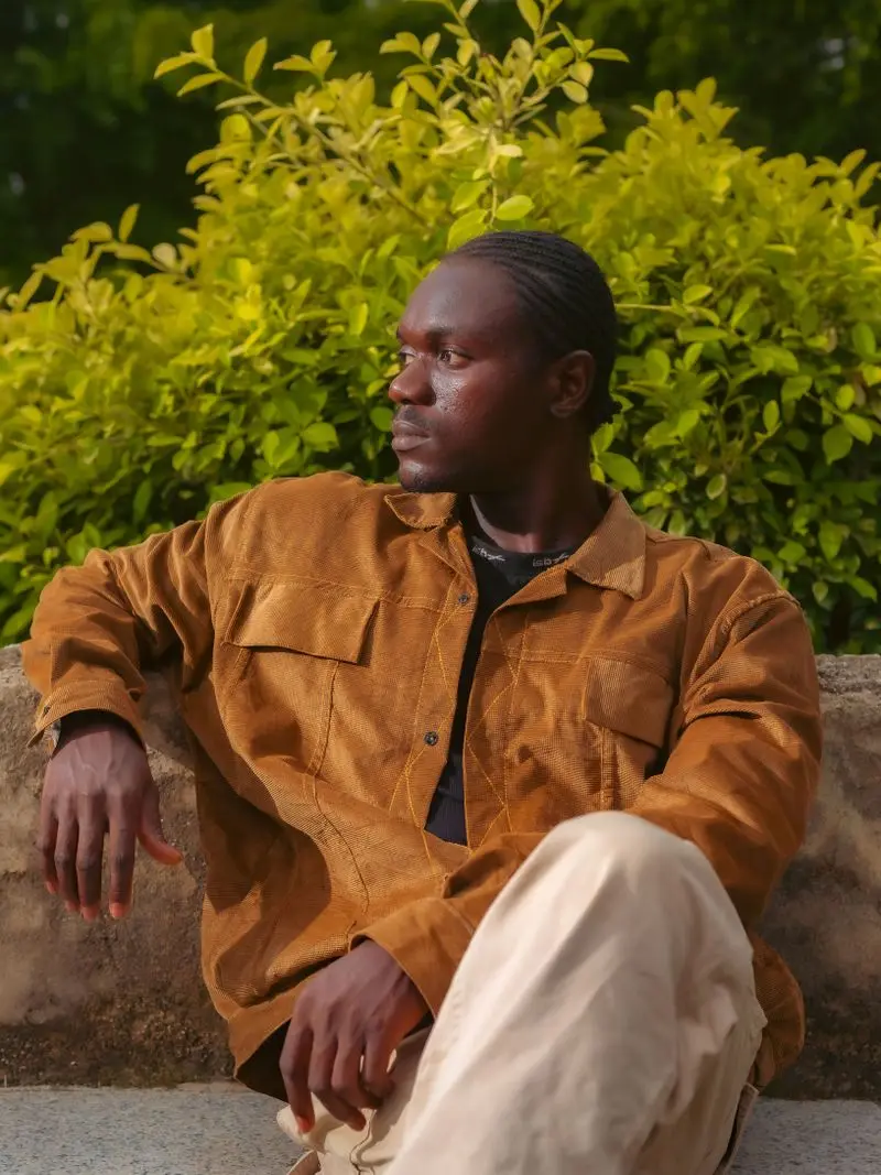 A man in a tan button-front jacket sitting outdoors against green leaves