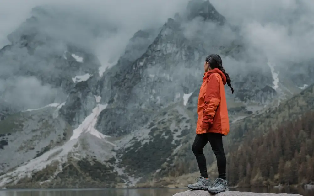 A hiker in an orange shell jacket standing on a rock below misty peaks