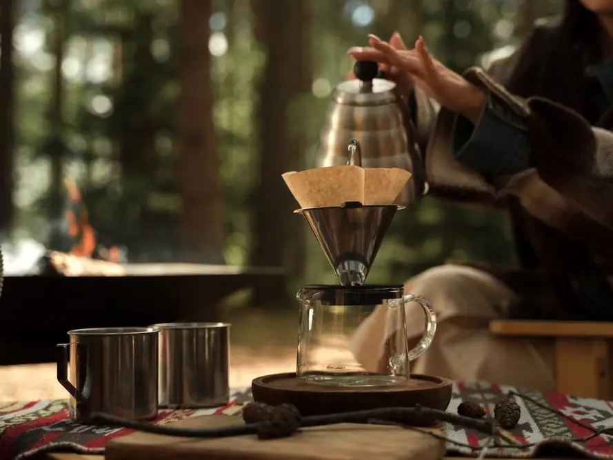 Hot water poured from a kettle into a pour-over coffee maker at an outdoor camp table